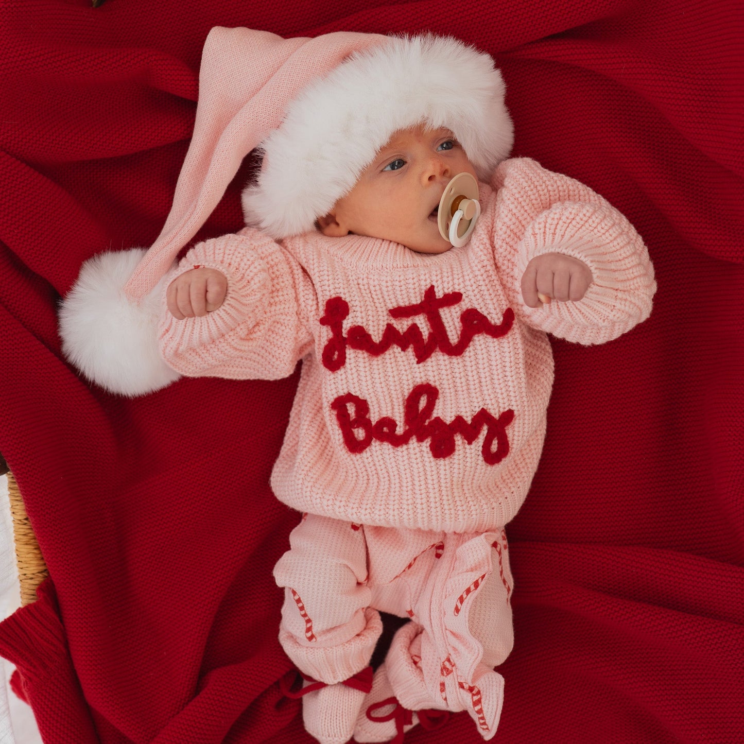 Baby in a pink 'Santa Baby' outfit with a white hat on a red blanket