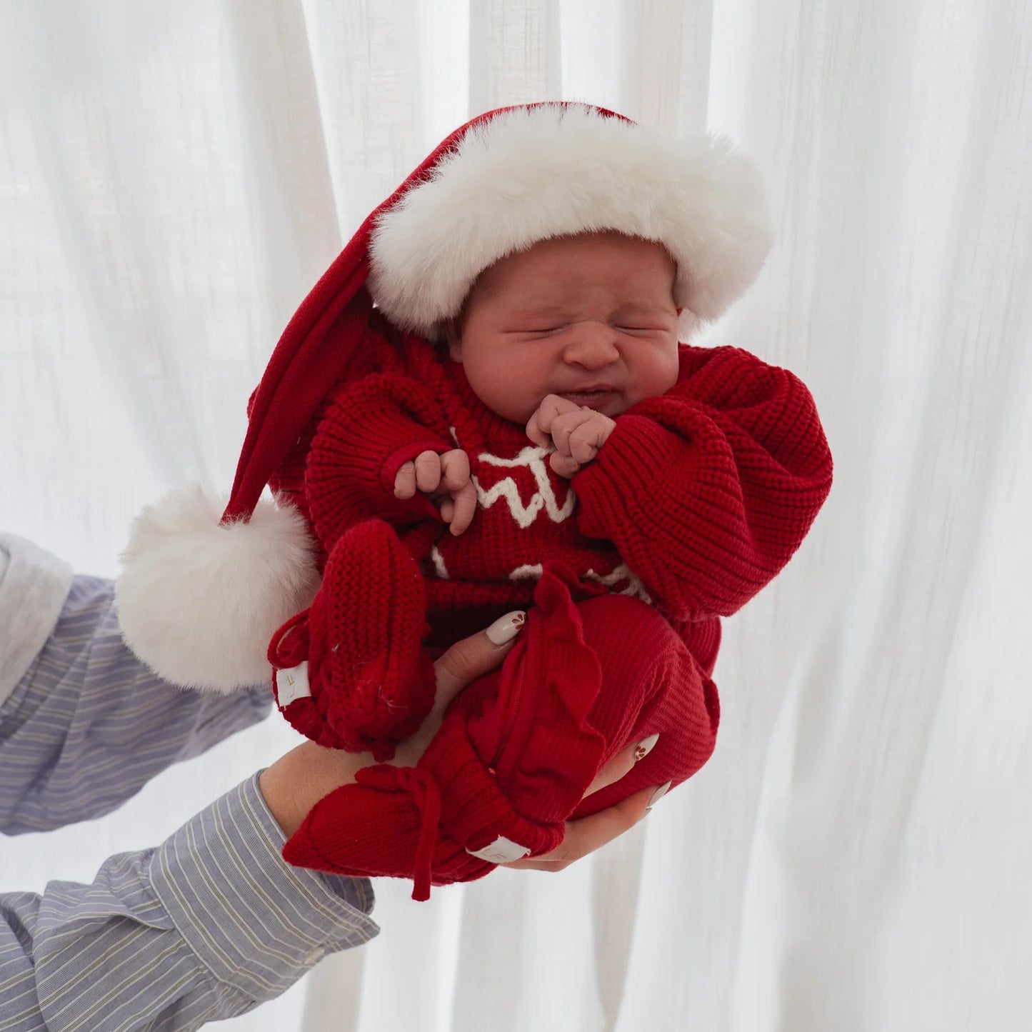 Newborn baby in a red outfit with white trim, held against a white curtain background.