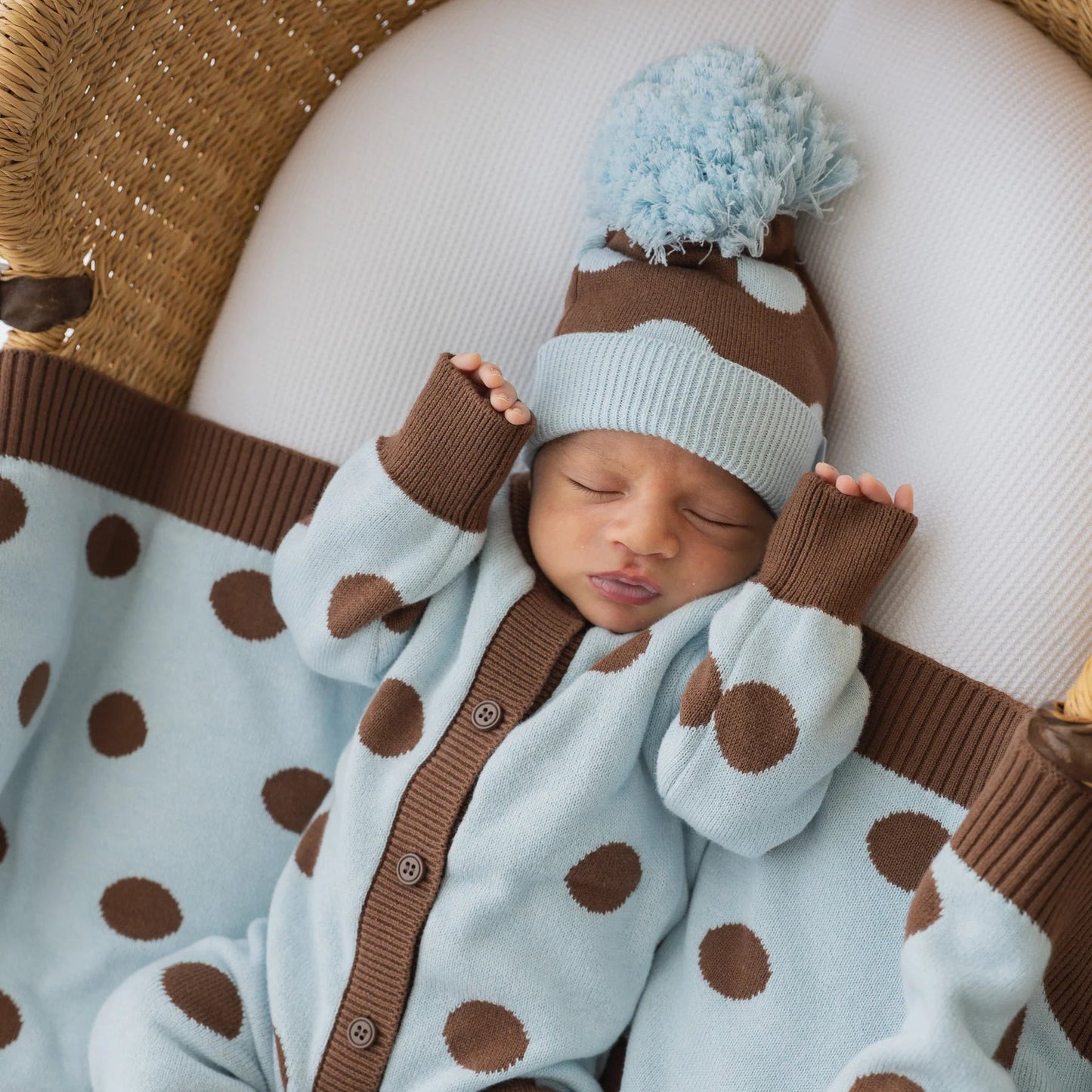 Newborn baby in a light blue and brown polka dot outfit with matching hat, lying on a textured surface.