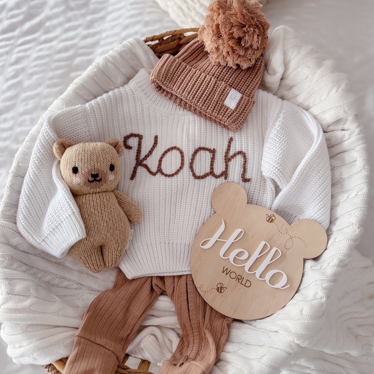 Baby outfit with personalised white sweater, brown pants, hat, and teddy bear on a white blanket.