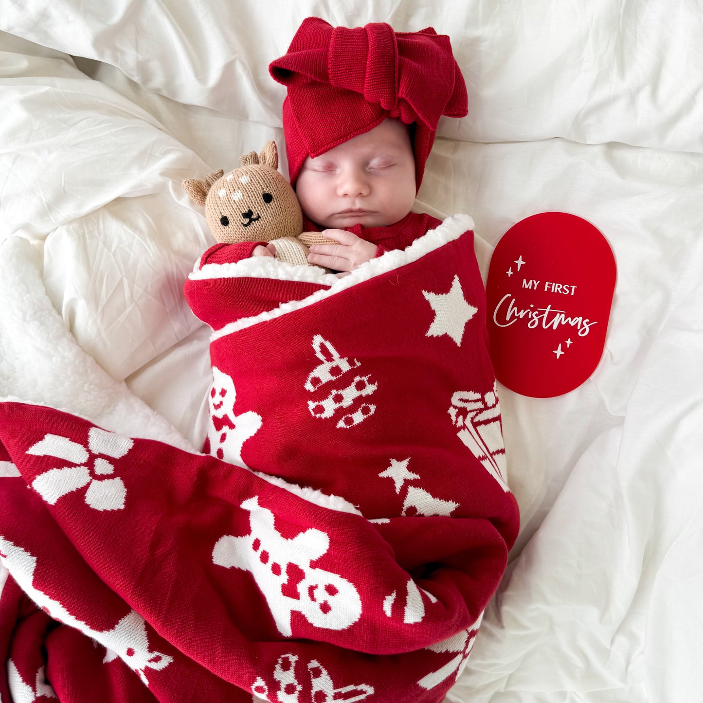 Newborn baby wrapped in a red blanket with white patterns, wearing a red hat and holding a reindeer teddy, on a white background.