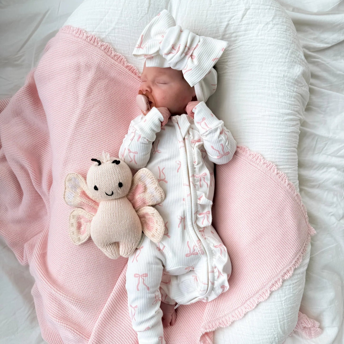 Newborn baby in a white outfit with a pink blanket and toy on a white surface