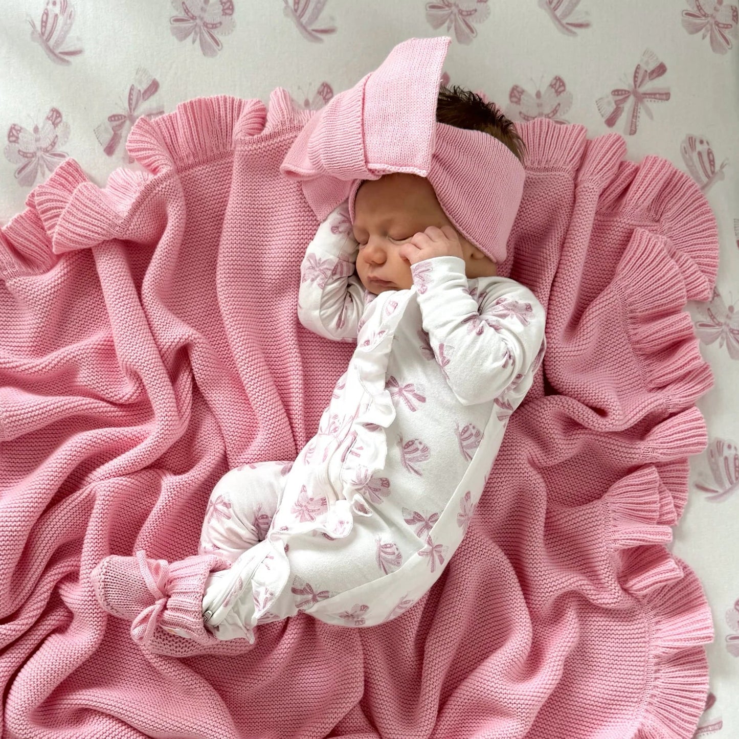 A newborn baby wearing a white and pink butterfly footed onesie and matching pink bonnet, sleeping on a pink ribbed blanket inside a crib with a pink patterned sheet.