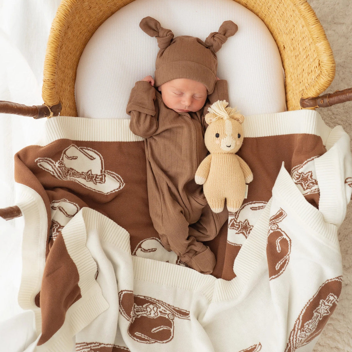 A newborn baby wearing a brown TEDDY hat and matching brown footed sleeper, lying in a woven rattan bassinet. The baby is wrapped in a brown and white cowboy hat print blanket and holds a small beige stuffed horse toy.