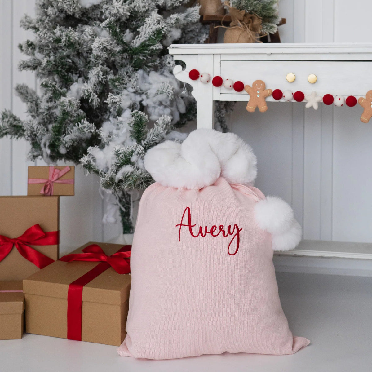 A light pink Christmas gift sack with fluffy white pom-poms and the name 'Avery' embroidered in red on the front, styled on the floor next to wrapped presents, a flocked Christmas tree, and a white mantel with holiday decorations.