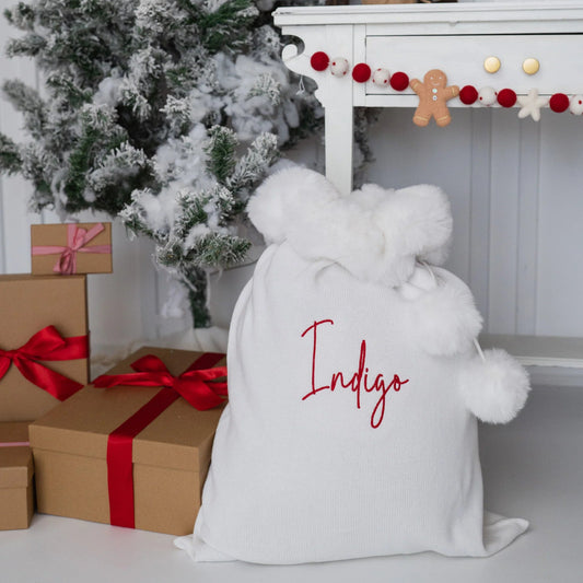 A white Christmas Santa sack with fluffy trim and the name embroidered in red on the front, styled in a festive holiday setting with wrapped gifts, a flocked Christmas tree, and a white mantel.