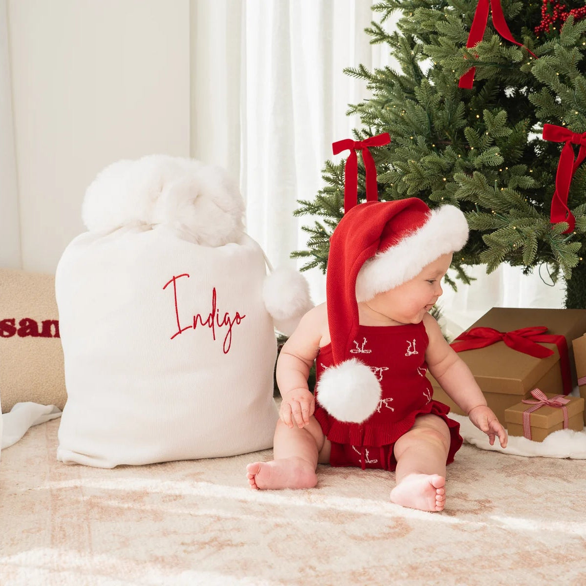 A baby wearing a red Christmas romper and Santa hat sits on a rug next to a white personalized Santa sack and a decorated Christmas tree with wrapped gifts.