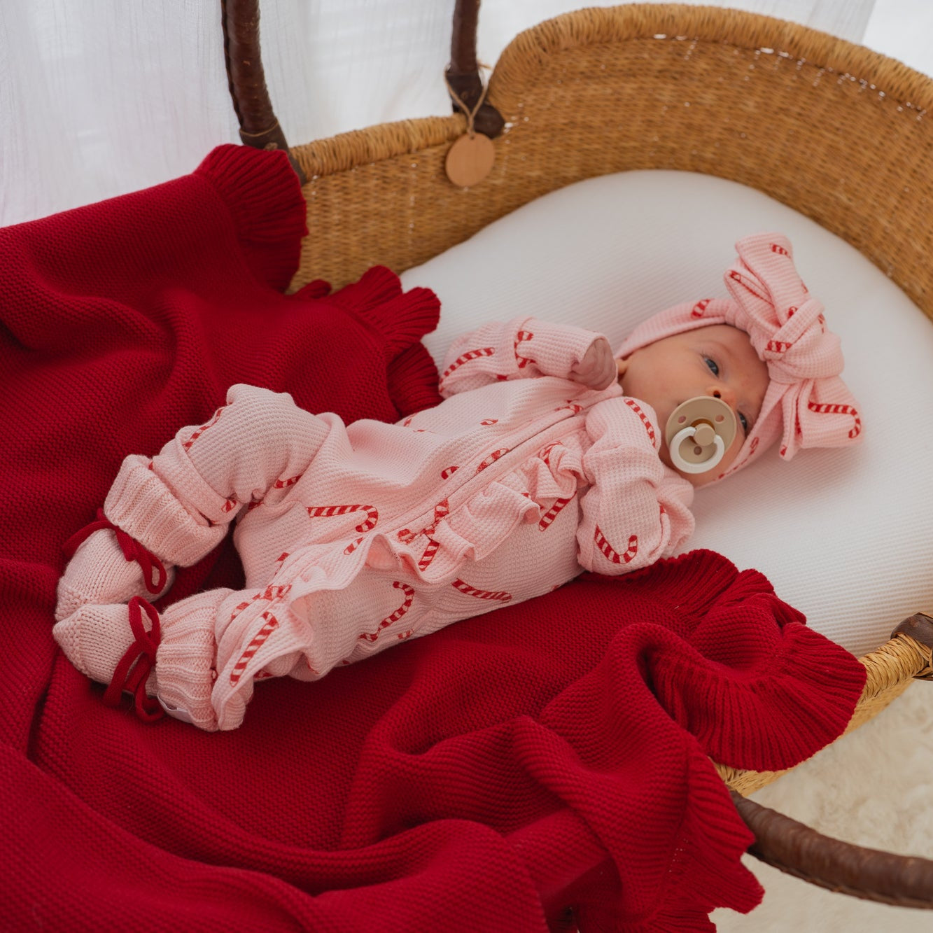 Newborn baby in a pink outfit with red accents lying on a red blanket inside a wicker basket.