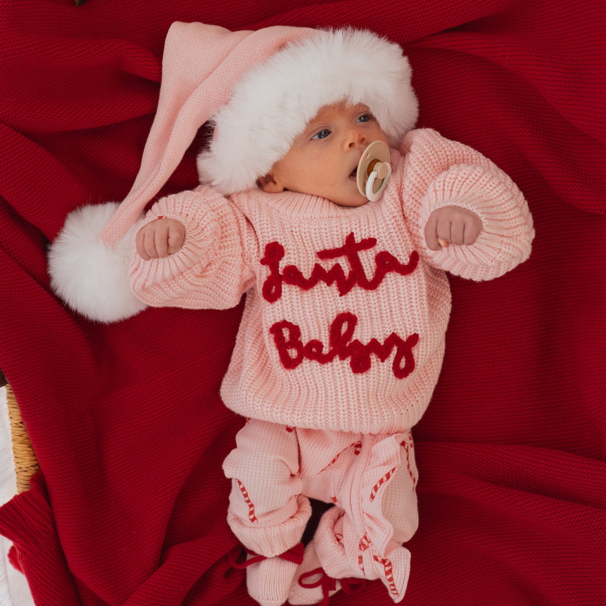 Baby in a pink 'Santa Baby' outfit with a white hat on a red blanket
