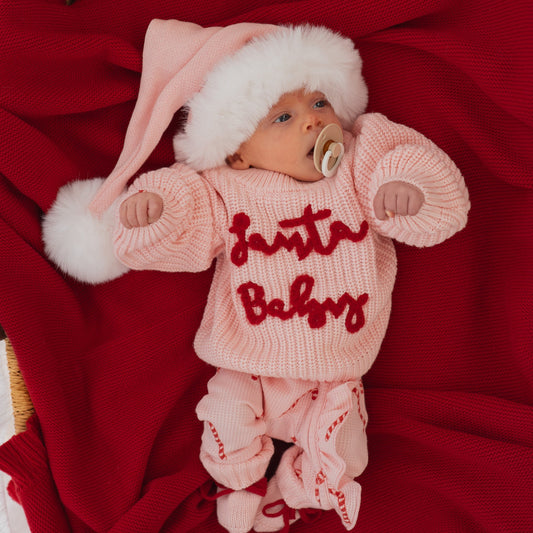 Baby in a pink 'Santa Baby' outfit with a white hat on a red blanket
