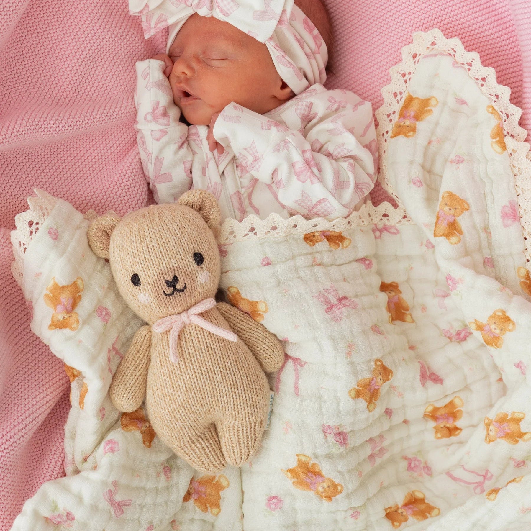 Newborn baby wrapped in a floral blanket with a teddy bear, wearing a matching headband on a pink background.