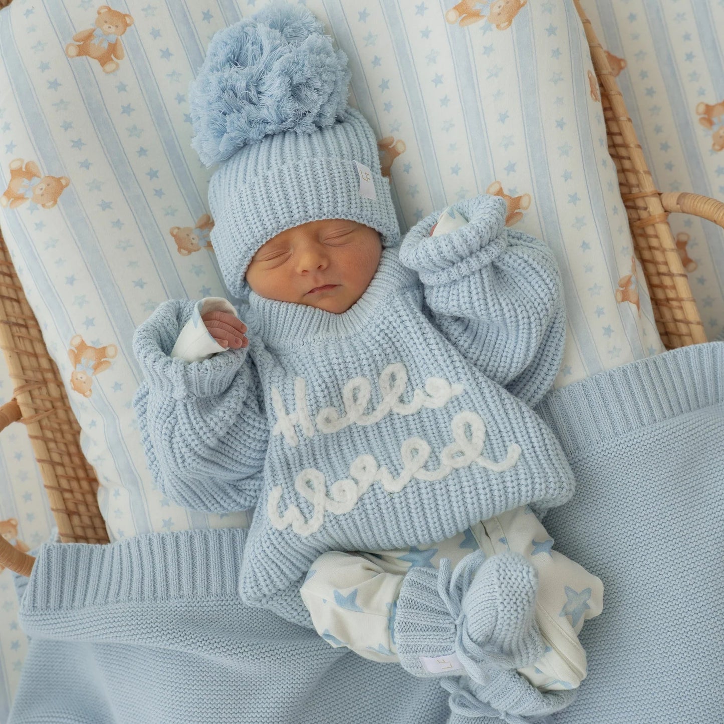 Newborn baby in a blue outfit with 'Hello World' text, wearing a matching hat, in a crib with a floral patterned sheet.