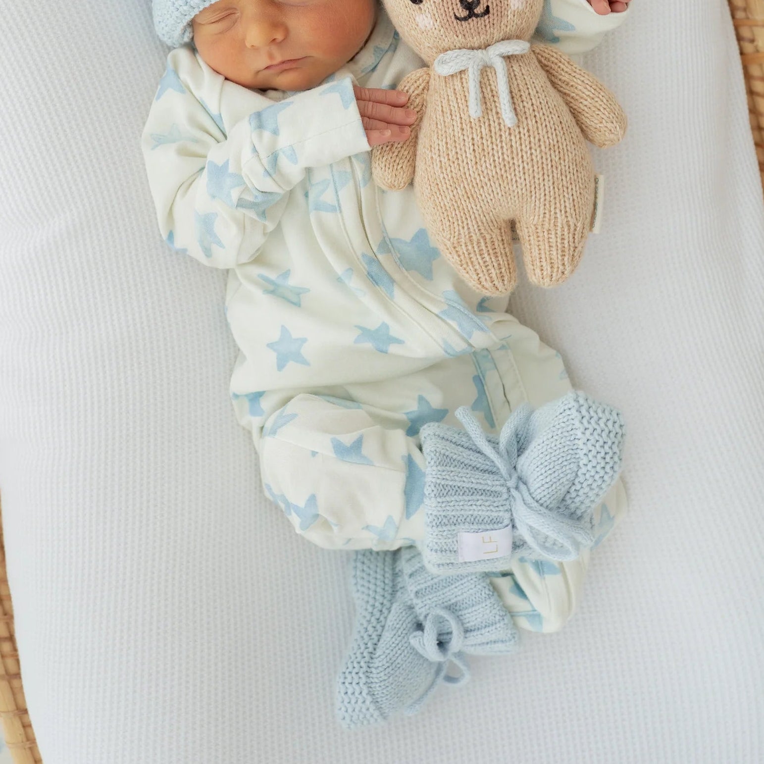 Newborn baby in a crib wearing a blue knitted BOOTIES and white outfit with star patterns, holding a teddy bear.