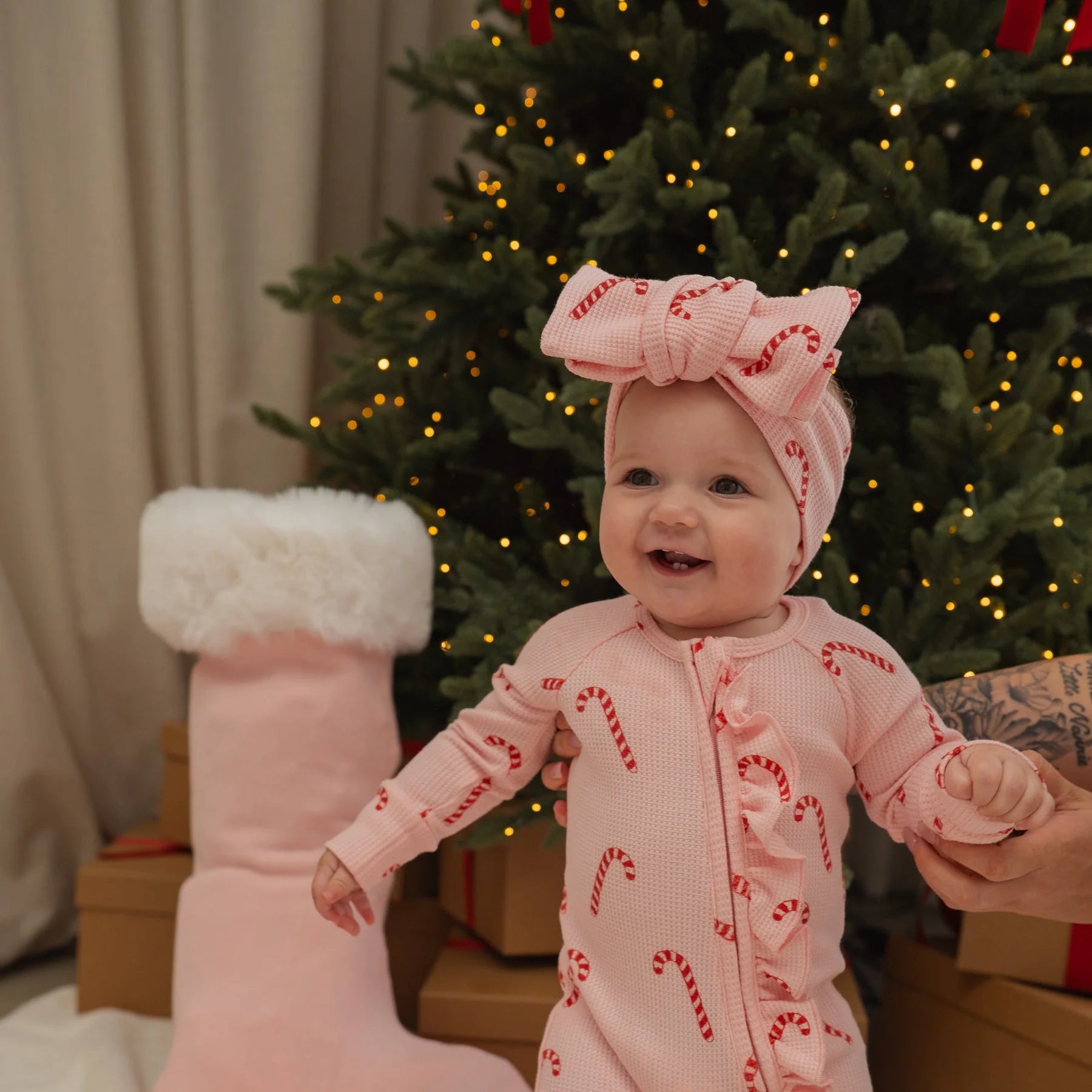 Baby in a pink outfit with candy cane pattern in front of a decorated Christmas tree.