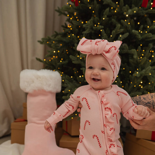 Baby in a pink outfit with candy cane pattern in front of a decorated Christmas tree.