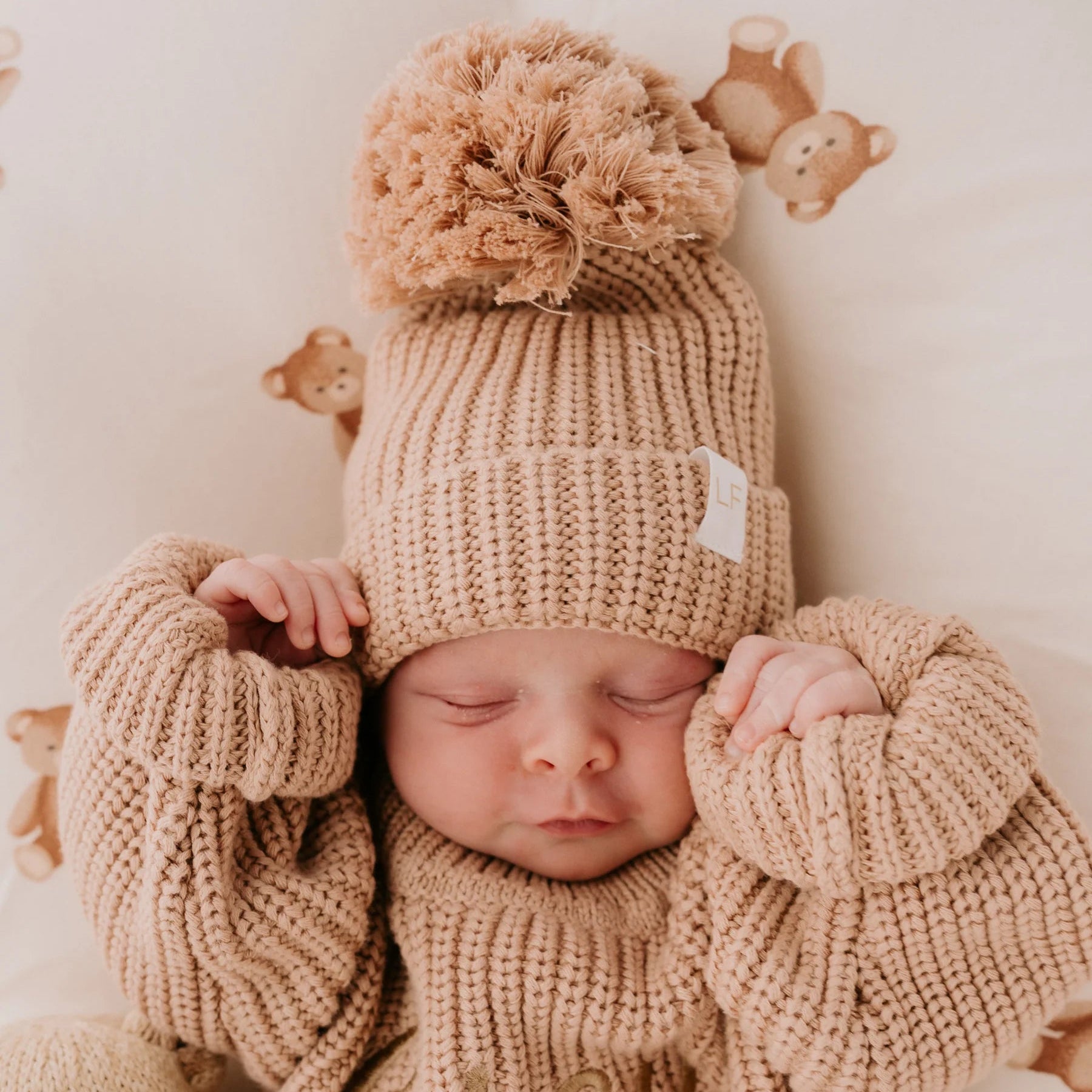 Baby in a beige knit outfit with a matching hat, surrounded by soft toys on a TEDDY PRINT SWADDLE.