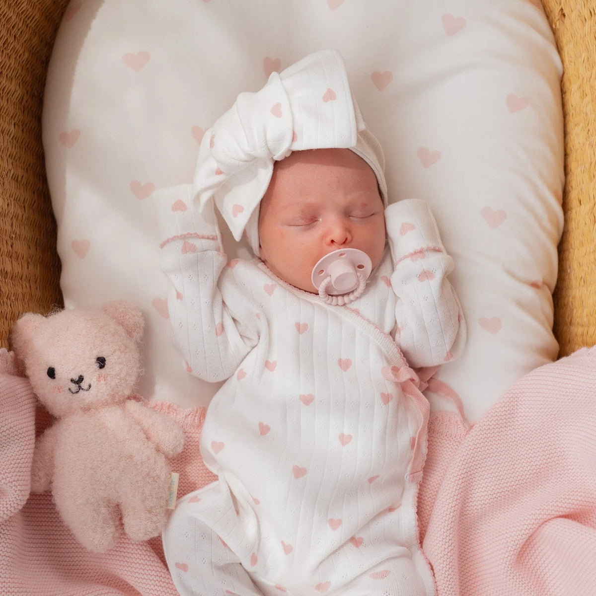 Newborn baby in a crib with pink blanket, teddy bear, and white outfit with pink hearts.