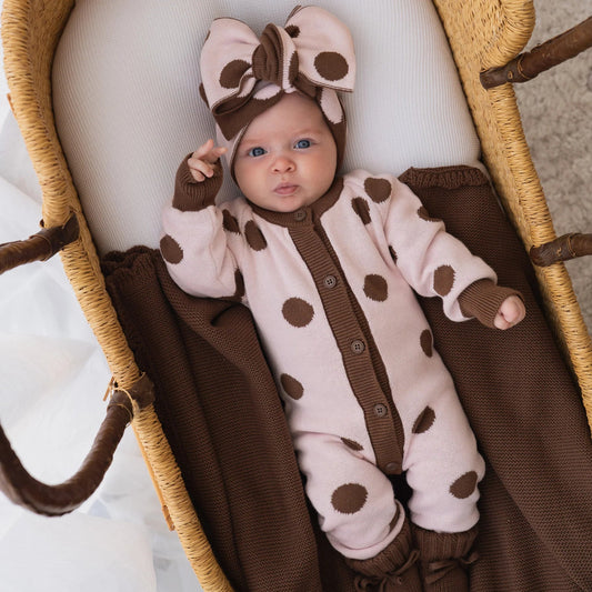 Baby girl in a brown and pink polka dot outfit with a bow, lying in a woven basket. Chocolate brown knitted blanket