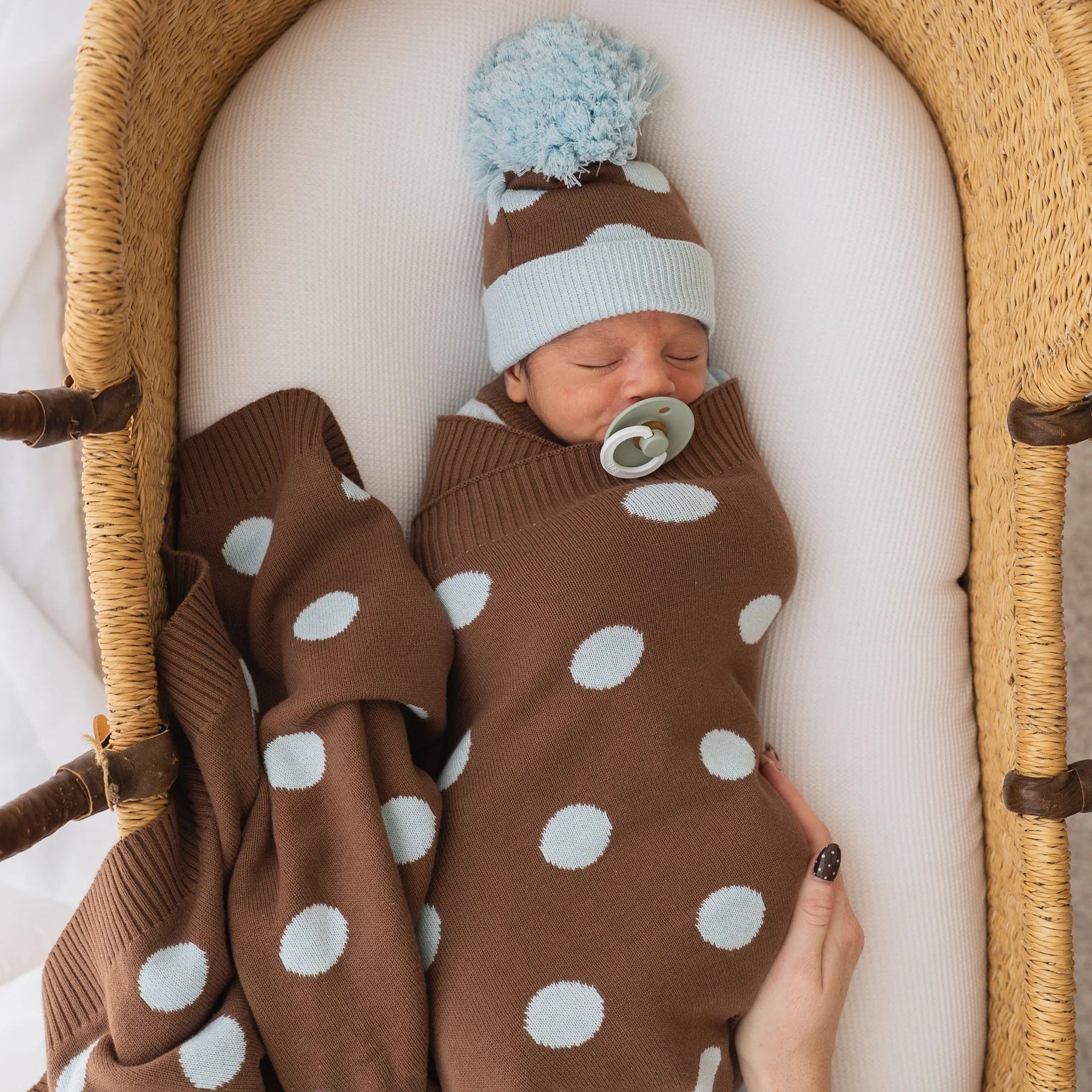 Newborn baby swaddled in a brown and white polka dot blanket with a blue and brown hat, lying in a wooden crib.