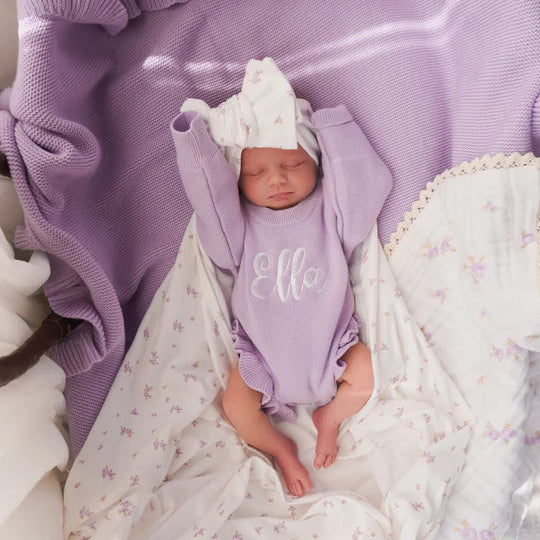 Newborn baby in a purple outfit with 'Ella' on it, wrapped in a purple blanket and lying on a white textured surface.