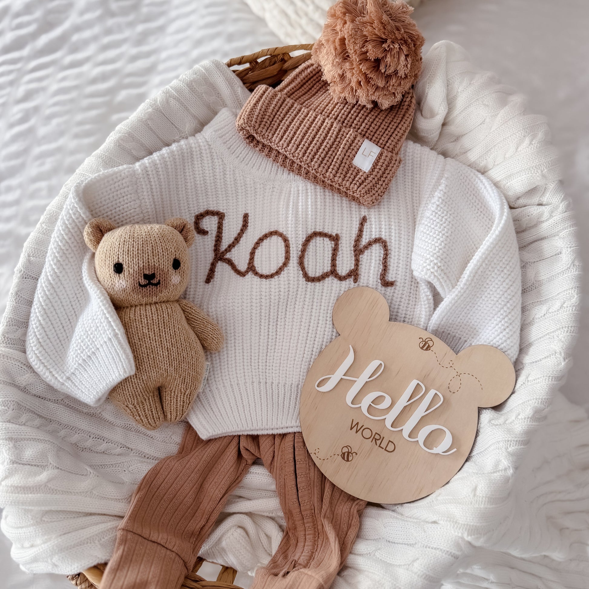 Baby outfit with personalised white sweater, brown pants, hat, and teddy bear on a white blanket.