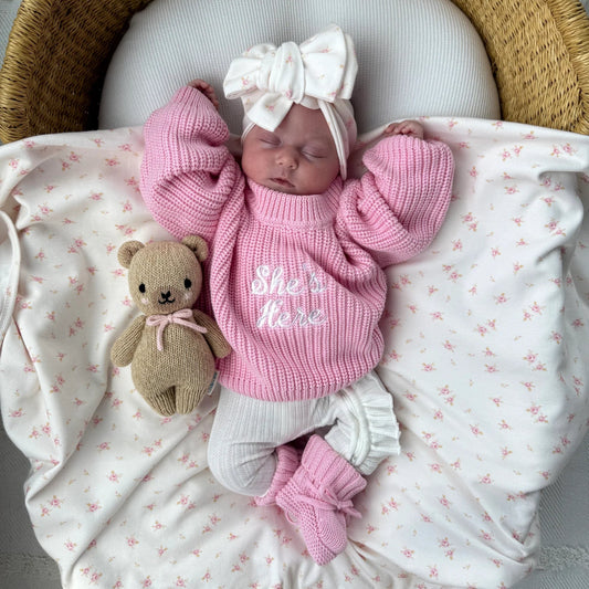 A newborn baby wearing a pink ribbed knit sweater with 'She's Here' in white script, a matching white bow headband, and pink booties, lying on a white blanket with pink star prints inside a woven wicker bassinet next to a small teddy bear.