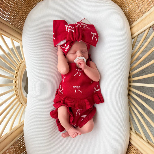 Newborn baby in a red outfit with a large bow, lying on a white pillow in a wicker basket.