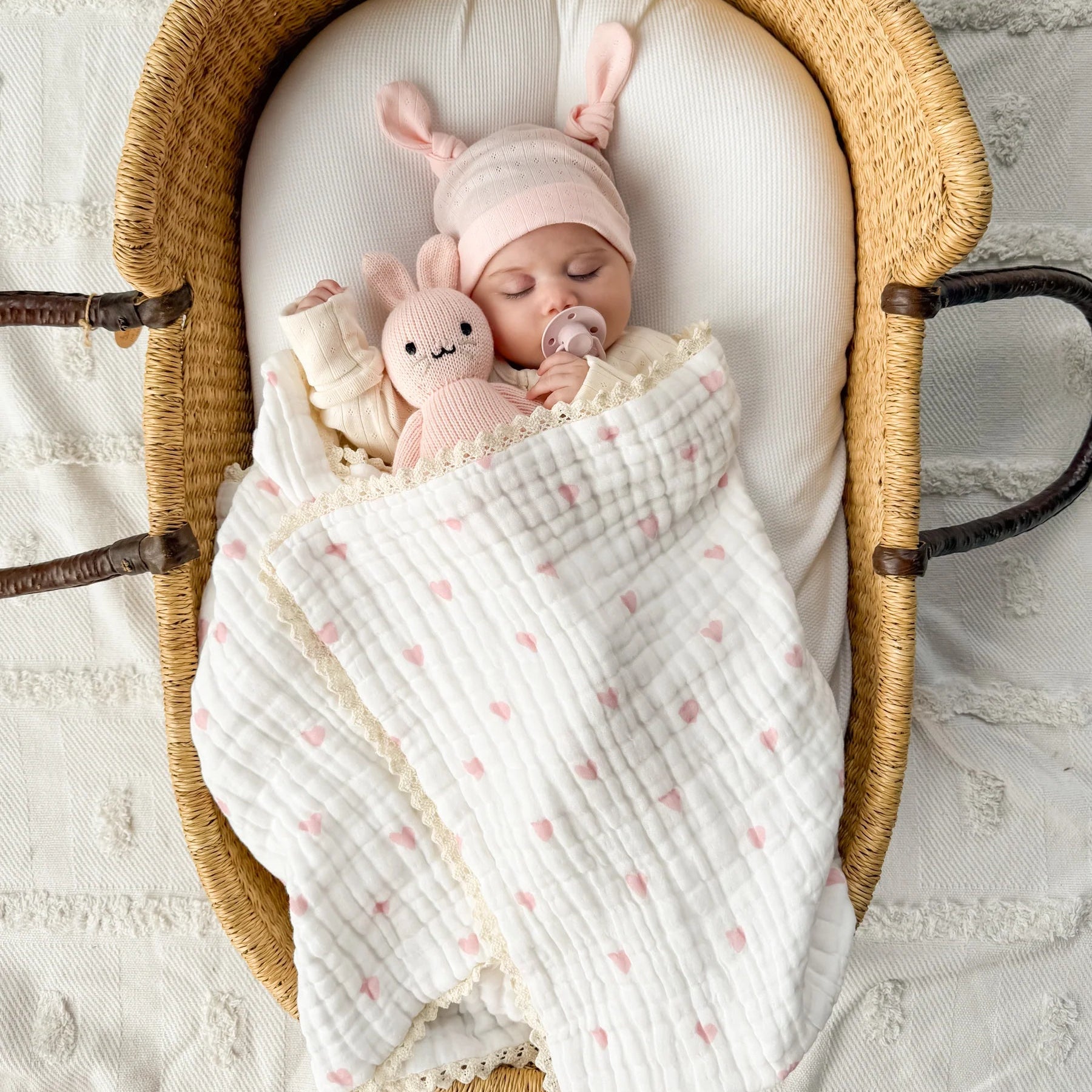 Baby in a Moses basket with pink bunny hat and plush toy, wrapped in a white blanket with pink hearts.