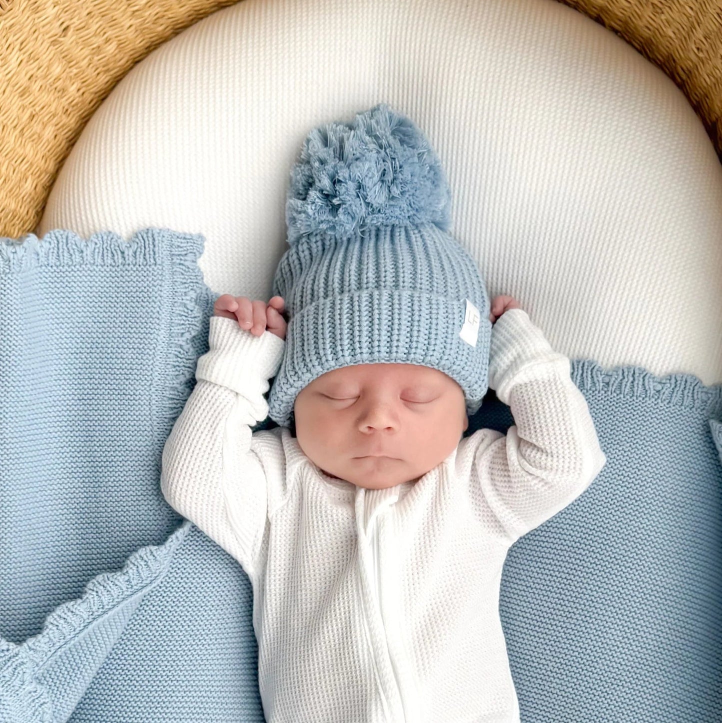 Newborn baby sleeping in a basket with a blue knitted hat and matching blanket.