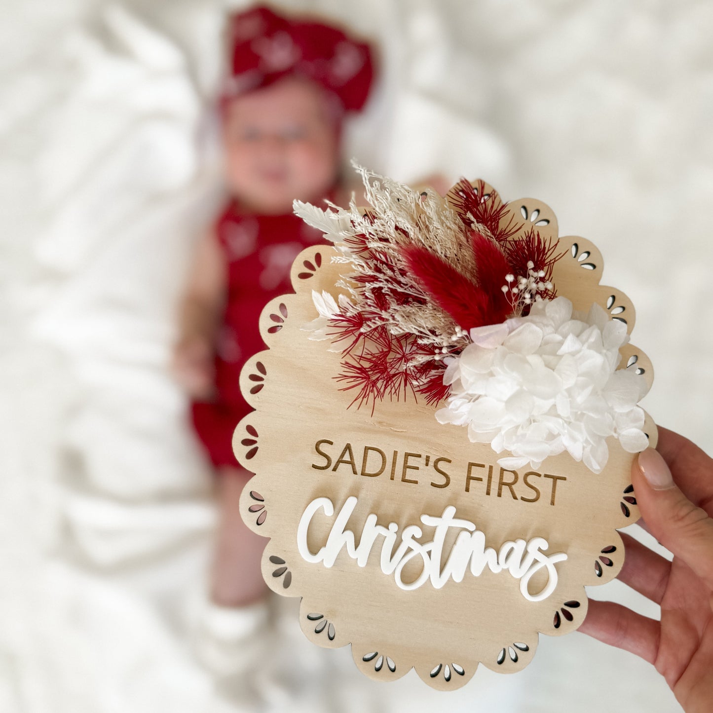 Wooden ornament with 'SADIE'S FIRST CHRISTMAS' text held by a hand, blurred background of a child in red.
