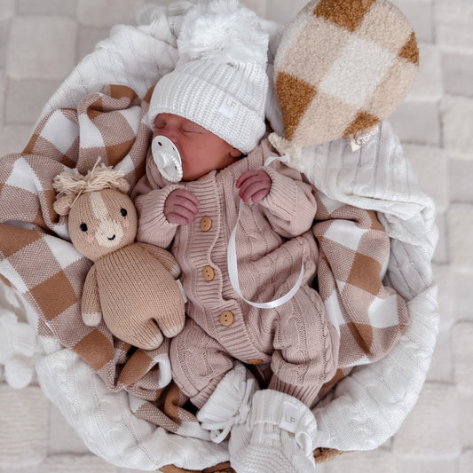 Newborn baby in a basket with checkered blanket, knitted cuddle and kind toy, and knitted beanie and booties, sleeping.