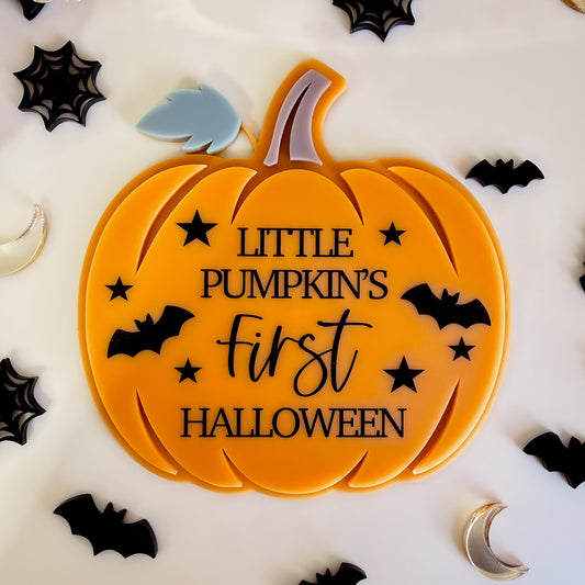 An orange pumpkin-shaped decorative sign with the text 'LITTLE PUMPKIN'S First HALLOWEEN' in black script and block lettering, surrounded by black bat and spider cutouts on a white surface.