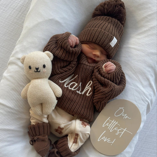 A newborn baby wearing a brown knit beanie with a pom-pom and a matching brown knit sweater with the name 'Nash' embroidered in white, lying on a white bassinet pillow next to a cream teddy bear toy and a round card that reads 'Our littlest love!'.