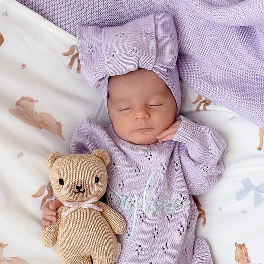 A sleeping newborn baby wearing a light purple knit onesie, matching bow headband, and booties, lying on a white horse-print blanket and holding a small beige teddy bear.