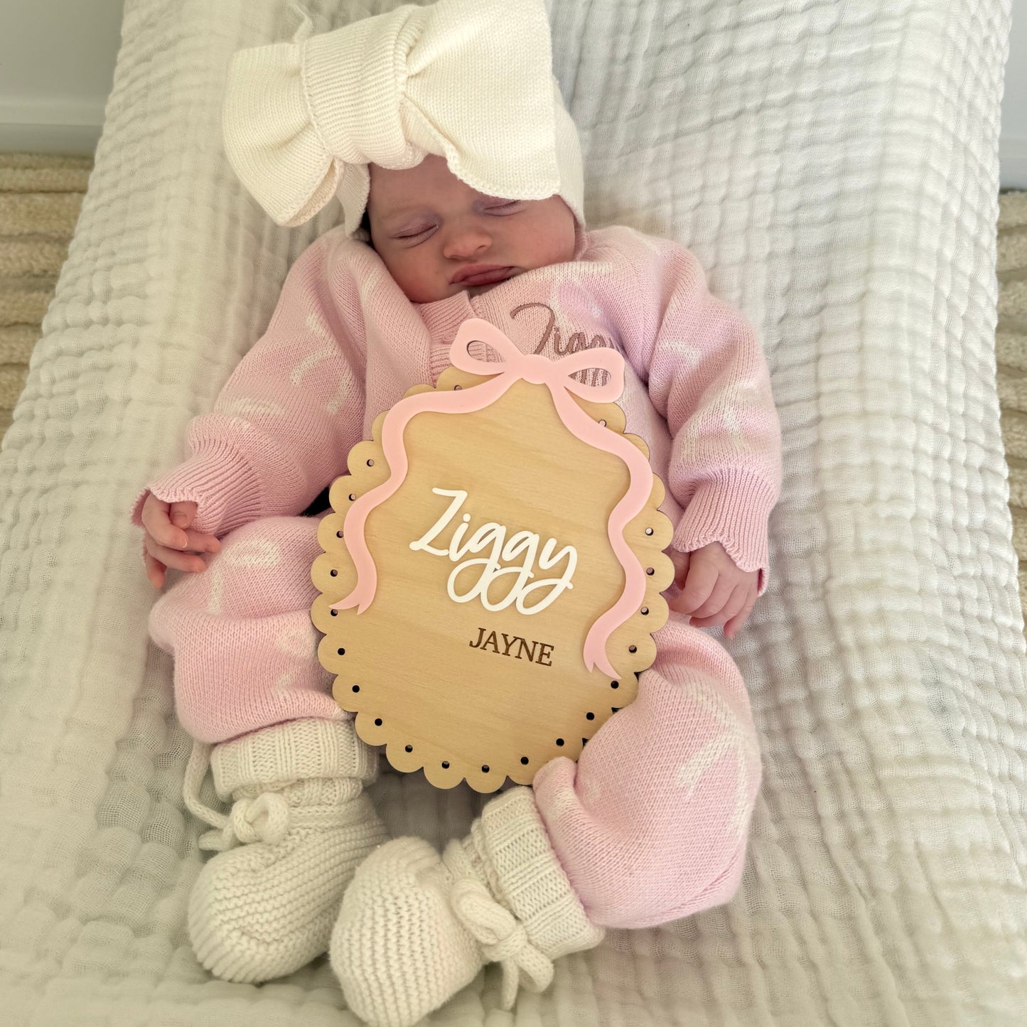 Newborn baby in pink outfit with a bow, holding a decorative sign with 'Ziggy Jayne' on a textured blanket.