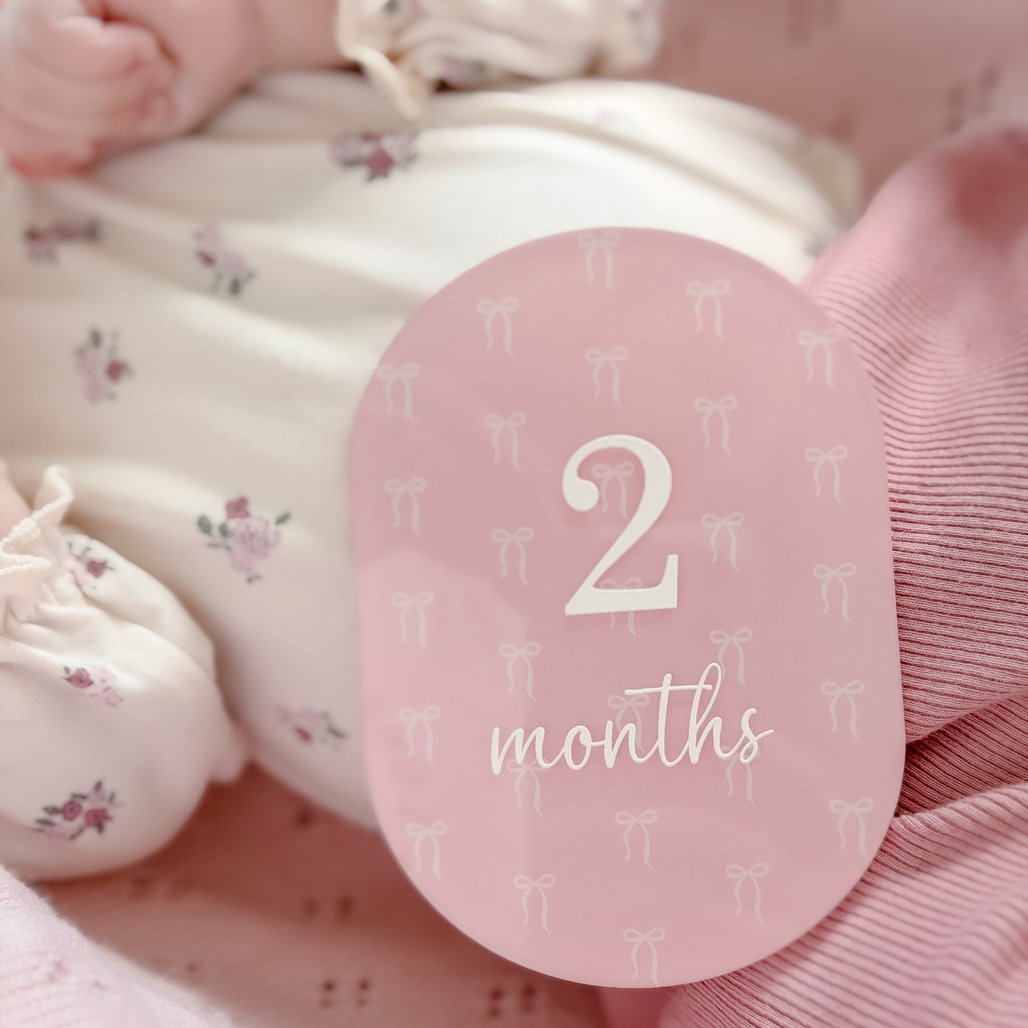 A close-up of a baby in soft pink and white floral clothing with a round pink milestone sticker that reads '2 months' resting on the baby's torso.
