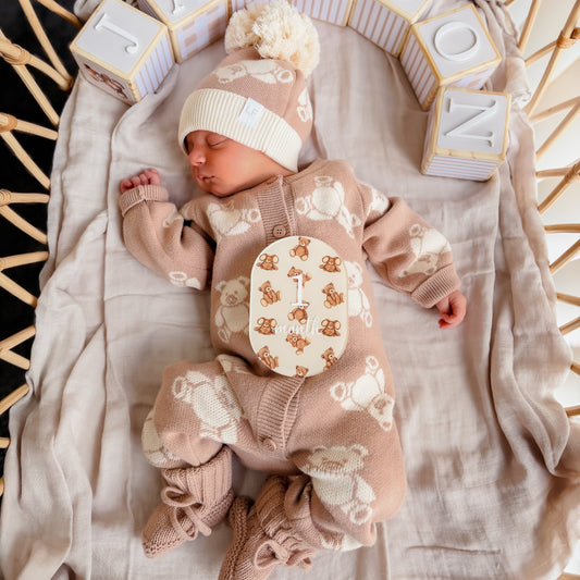 A sleeping newborn baby wearing a beige and cream teddy bear patterned knit onesie, matching beanie with pom-pom, and booties, lying on a muslin blanket inside a wicker basket. Wooden letter blocks spelling 'JACKSON' are arranged behind the baby.